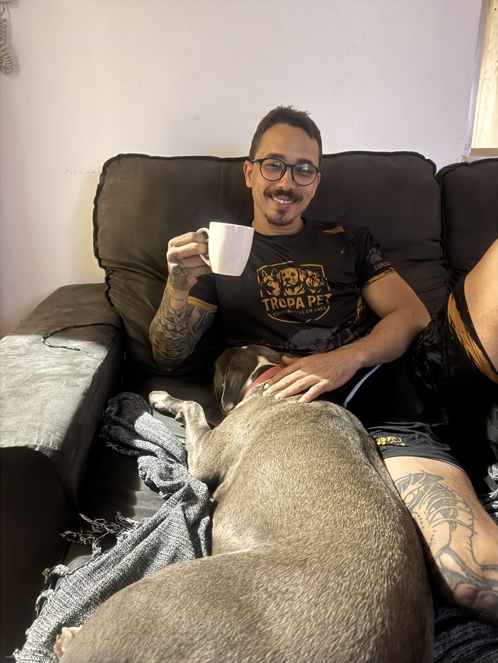 Jordan sorrindo no sofá com uma xícara de café, com a cachorra hospedada deitada no colo